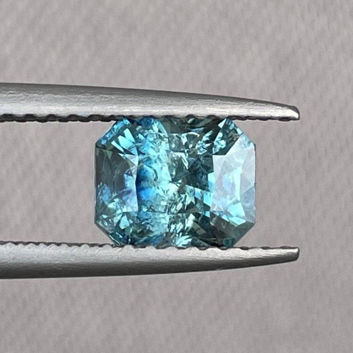 Teal sapphire gemstone from NASHGEMS, radiant cut, 5.40 x 6.50 mm, 1.38 carats, showcasing vibrant blue-green color and clarity.